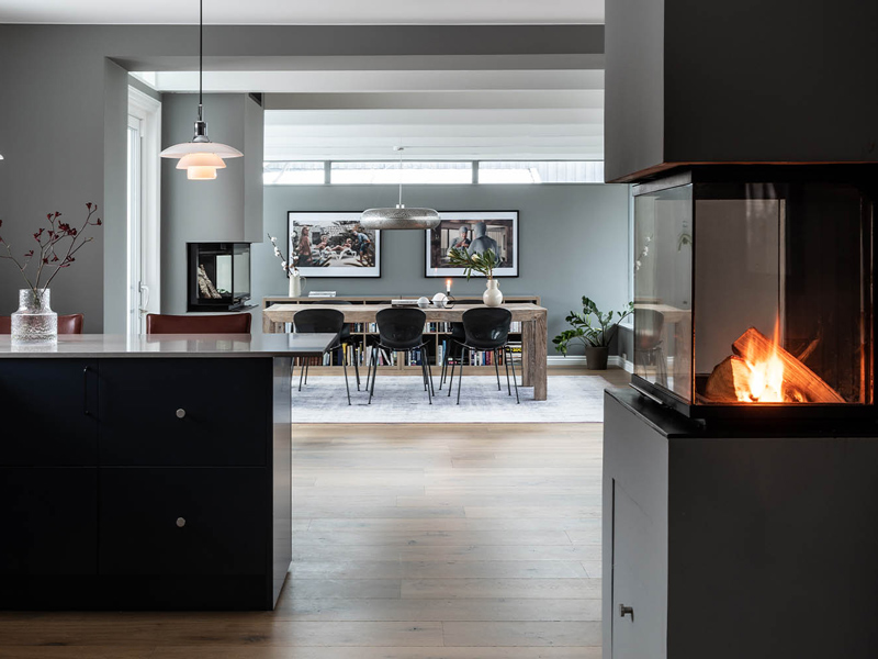 Bildet viser en lekker leilighet med stilfullt interiør. Det er et kjøkkenet med svarte fronter i forkant, en peis lyser opp til høyre i bildet og man ser ut mot spisestuen i bakgrunnen.