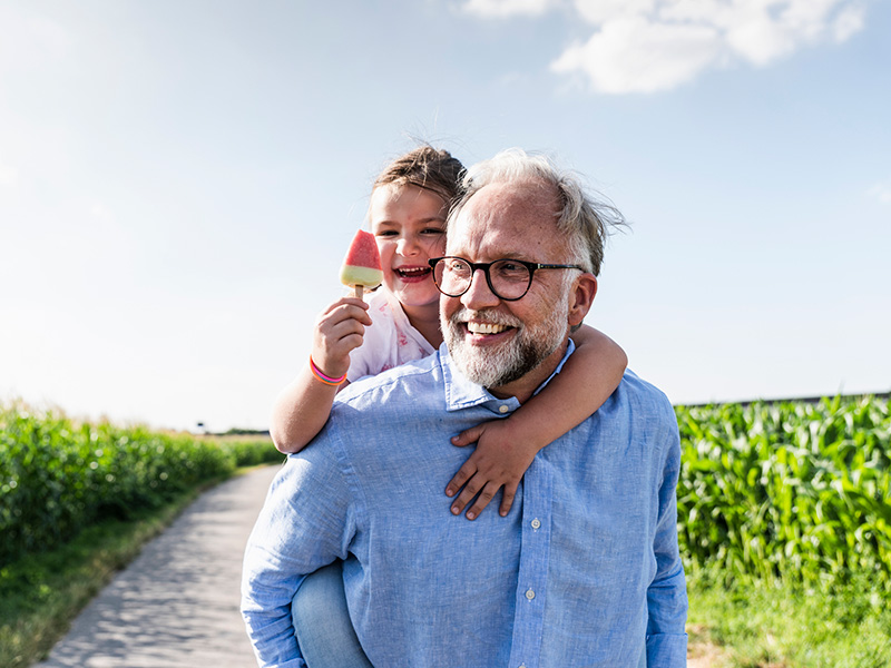 Bilde av en Bestefar med barnebarn på skuldrene ute på tur i grønne omgivelser.
