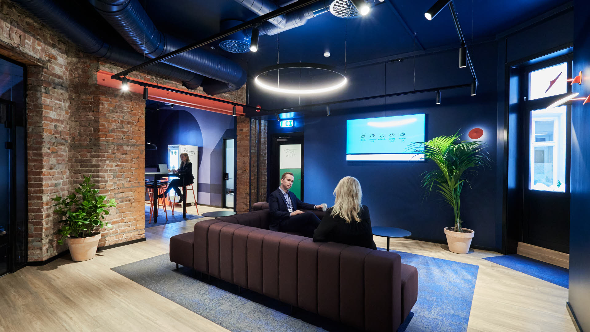 Bildet viser fellesområdet i Thon Flex lokalet i Karl Johans gate 5. To mennesker snakker sammen i en sofa. Sofaen er brun, blått teppe. Veggene i lokalet er delvis malt i blått og noen elementer med murstein på motsatt vegg.