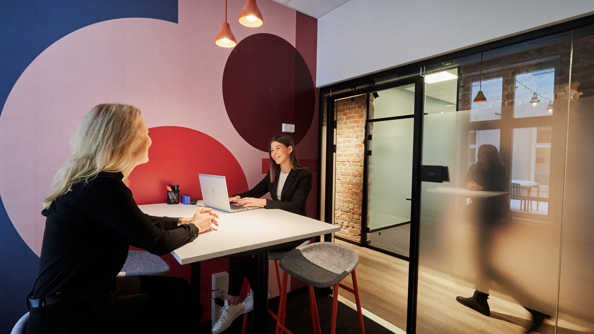 Bilde av møterom i Thon Flex, Karl Johans gate 5. To damer prater sammen i et lokale som har store glassvinduer, høye stoler og en lilla vegg med oransje maleri.