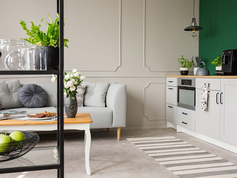 Bilde av et hvitt kjokken med mørk grønn farge på veggen bak. Grå sofa skimtes bak en svart hylle. Grønne planter og hvite roser gjør rommet ekstra hyggelig.