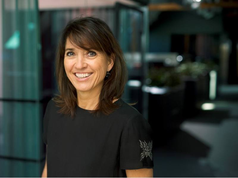 Dame med mørkt hår og mørke klær ser smilende inn  i kamera, mørkt rom med glassvegger i bakgrunnen. Foto til artikkel om boligtrender.