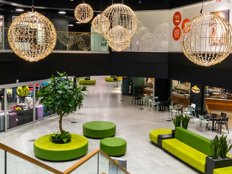 Bilde fra kjøpesenteret AMFI Roseby. Viser fellesområdet med grønne benker, et grønt kunstig tre og flotte lysekroner i taket.